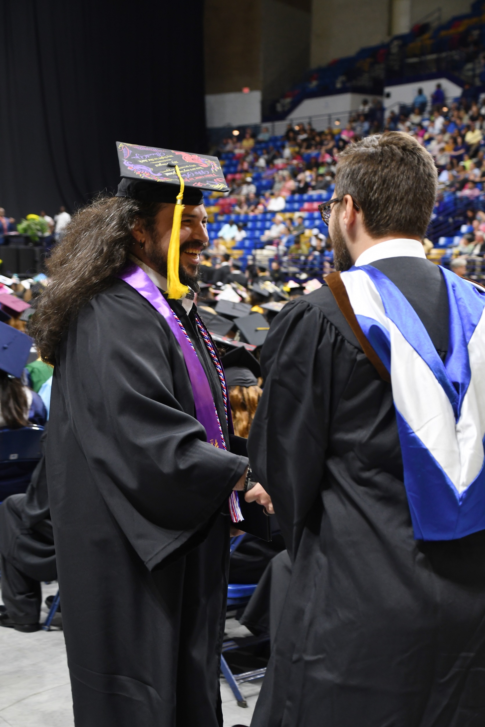A graduate with long hair and a beard and wearing a purple stole shakes hands with a faculty member. 