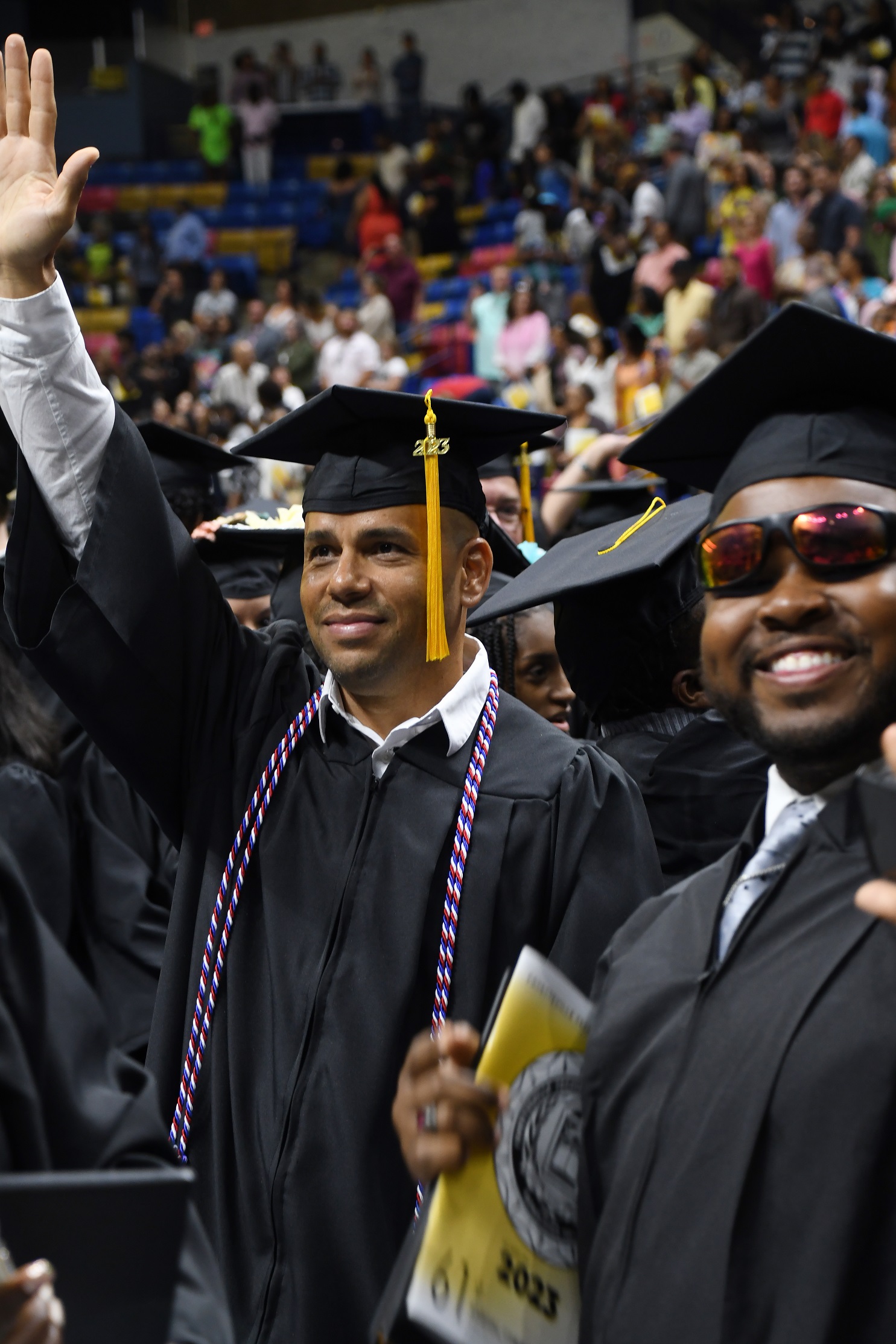 A graduate wearing a red, white and blue cord waves as someone in the crowd.