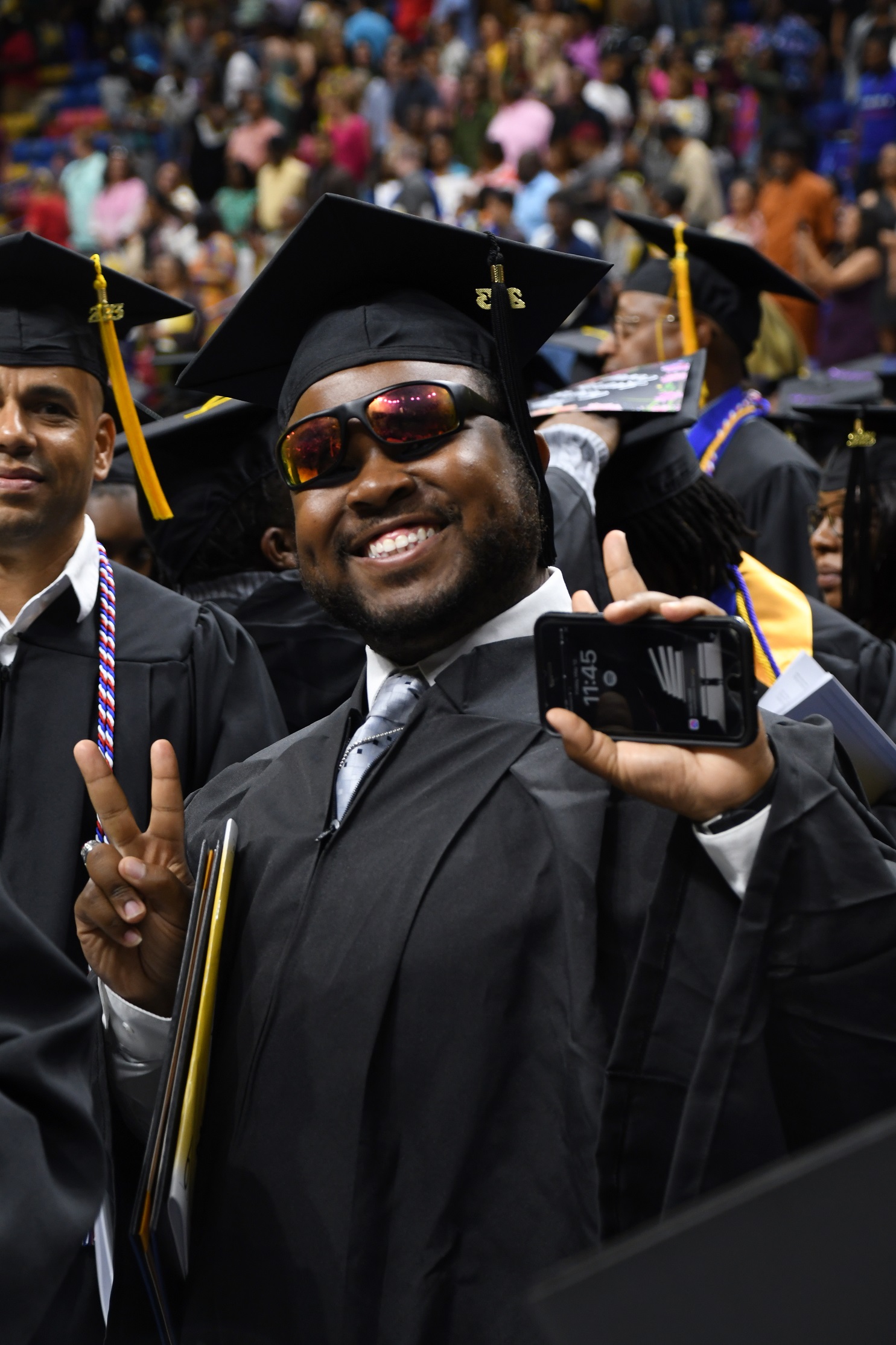 A graduate wearing sunglasses smiles and holds up two fingers in a "peace sign."