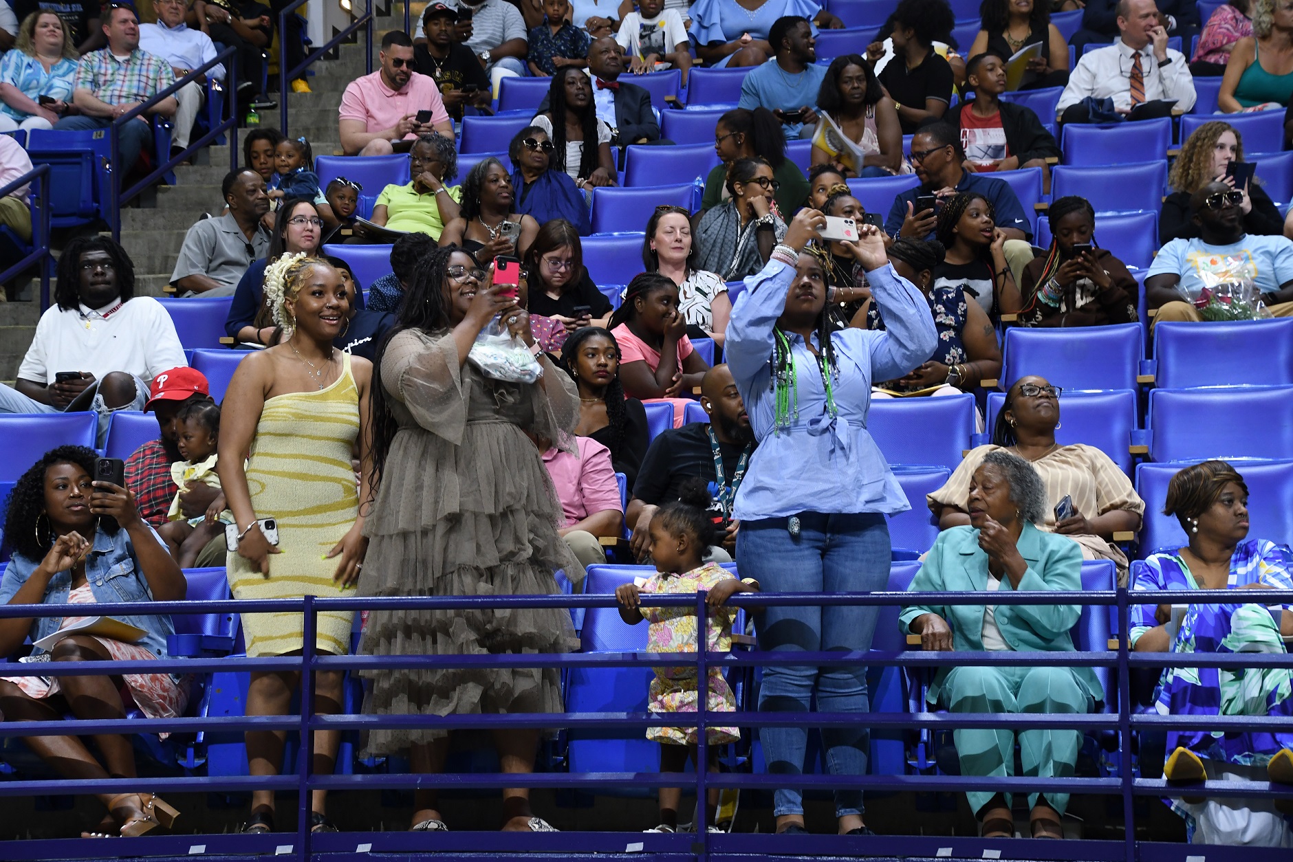 Three woman stand at the railing at the front of the stands. Two of them have their phones up to take pictures.