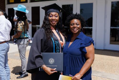 A graduate and a woman pose together for a photo. The graduate is holding her diploma folder.