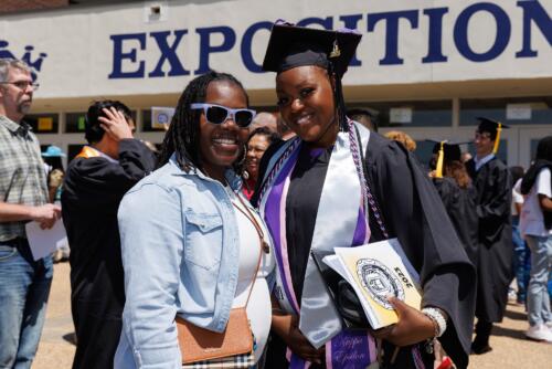 A graduate wearing several stoles and cords poses with a woman for a photo in front of the Crown Expo doors.
