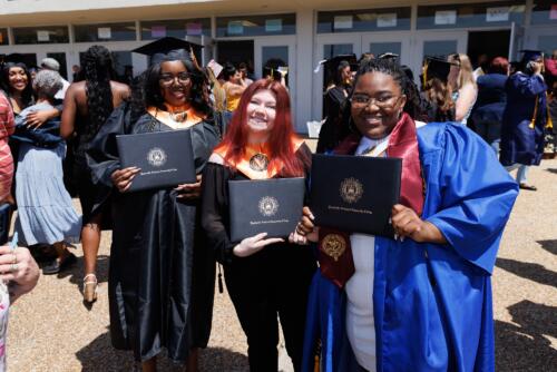 Three graduates lean together and hold up their diploma trophies to pose for a photo.