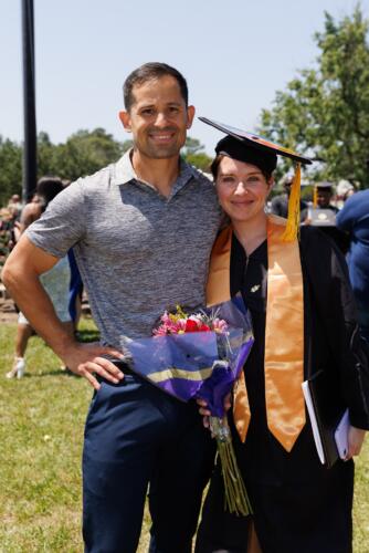 A graduate in her cap, gown and stole, holds a bouquet of flowers as she poses for a photo with a man.