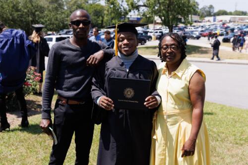 A graduate holds up his diploma folder while he stands between a man and woman outside the Crown.