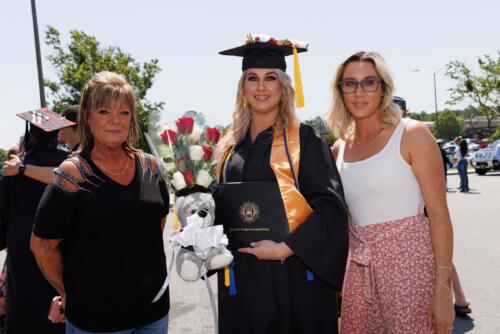 Three woman pose together. The one in the middle is dressed in her cap, gown, and stole and holding her diploma folder.
