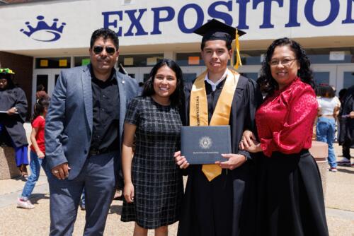 A graduate wearing his cap, gown, and stole and holding his diploma folder poses with his family.