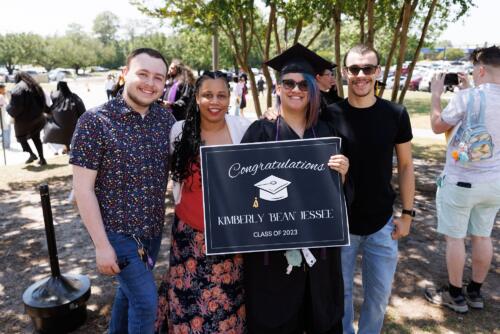 A graduate stands with her loved ones. She's holding a sign that says "Congratulations Kimberly Bean Jessee. Class of 2023."
