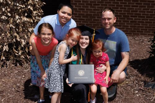 A graduate kneels down next to three children. Two adults kneel behind them. The graduate is holding her diploma folder. 