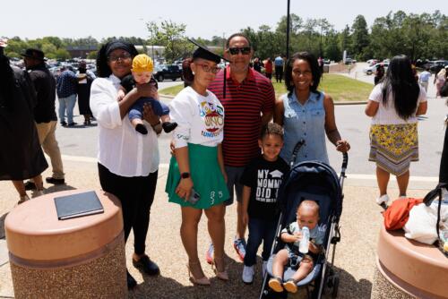 A graduate, who is wearing her cap with an outfit of a sweatshirt and skirt, stands with her loved ones outside of the Crown.