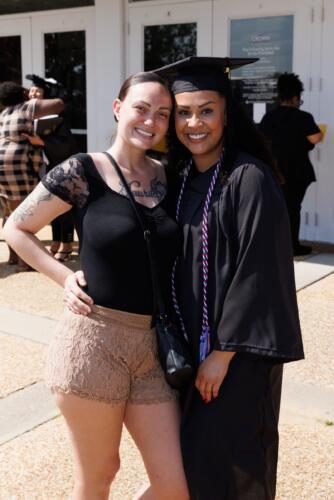A graduate wearing her cap, gown and a red, white and blue cord, poses with a woman outside of the Crown.