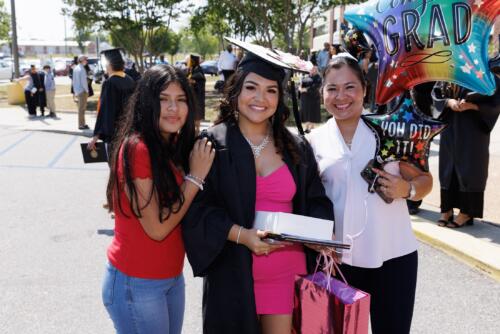 A graduate wearing her cap and gown and holding a gift bag poses with two women outside of the Crown.