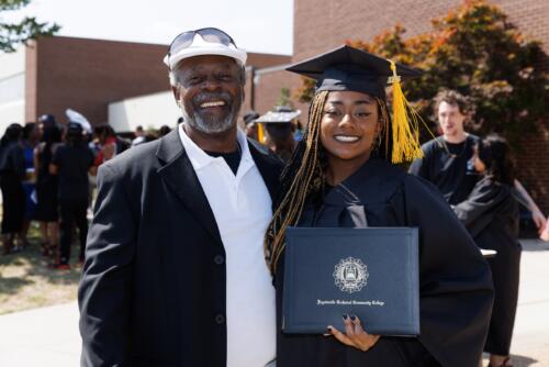 A graduate wearing her cap and gown and holding her diploma folder poses for a photo with a man in a white shirt and dark blazer.