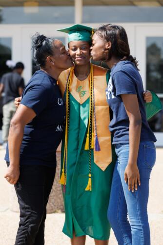 Two women lean in to give a kiss on either cheek to a graduate in a green cap and gown and gold stole.