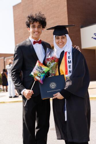 A graduate wearing a cap and gown and a stole that says "Yemen" poses with a man in a suit and bowtie.