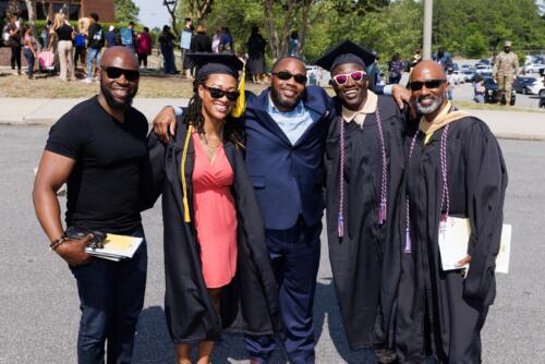 Three graduates pose with loved ones outside. They are all wearing sunglasses.