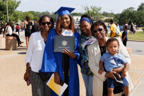 A graduate wearing a blue cap and gown and a silver stole poses with her loved ones.