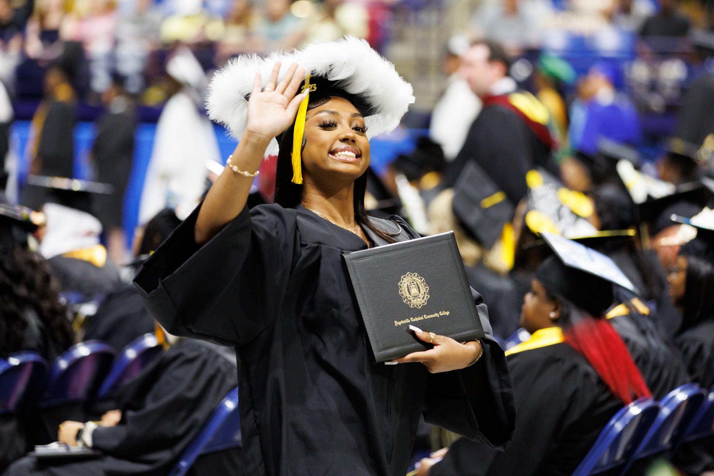 A graduate with a decorated cap waves to the crowd while holding her diploma folder.