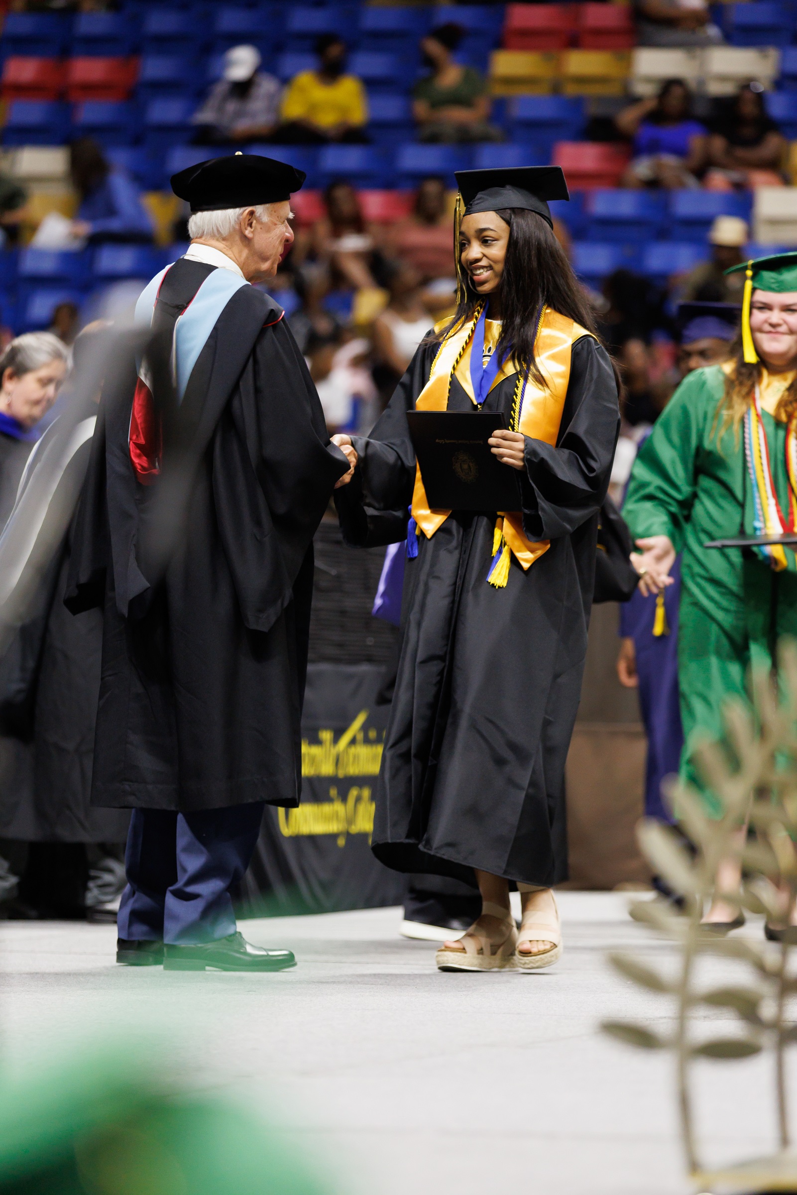 A graduate wearing a gold stole  and a medallion on a blue ribbon shakes hands with Charles Koonce as she walks across the stage.
