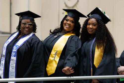 Three graduates, each wearing a personalized stole, wait in line in the staging area.