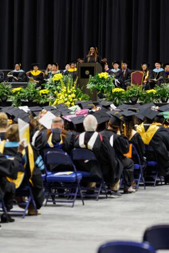 A photo of Tammy Thurman speaking at the podium on stage. Rows of faculty members, shown from behind, are visible in the foreground.