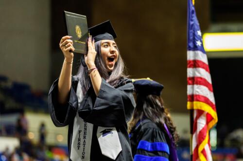 A graduate wearing a white stole that says "First Generation" holds up her diploma folder in celebration.