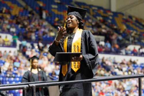 A graduate wearing a gold stole that says "Student Athlete" raises her hand to her mouth in emotion after receiving her diploma folder.