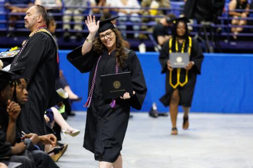 A graduate wearing a red, white and blue cord waves and holds her diploma folder while walking back to her seat.