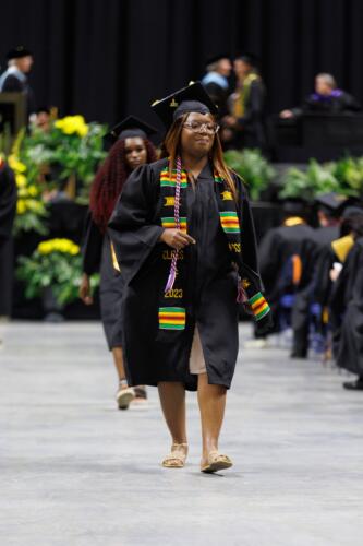 A graduate wearing a red, white and blue cord and a black, green, red and yellow stole walks back to her seat.