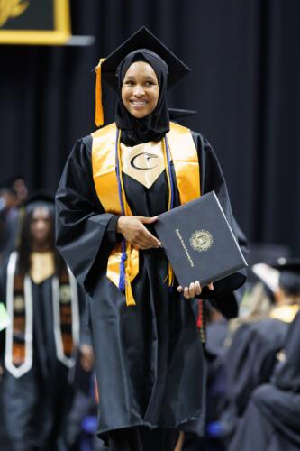 A graduate wearing a gold stole with a hawk logo on it smiles as she walks back to her seat. She is carrying her diploma folder.