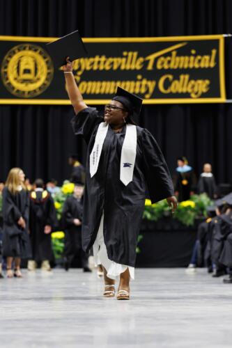 A graduate wearing a white stole that says "First Generation" on it lifts her hand to wave at the crowd.