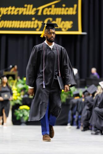 A bearded graduate wearing a red, white and blue cord walks back to his seat while carrying his diploma folder. 