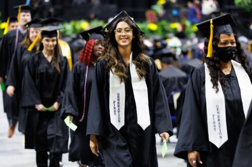 A graduate wearing a white stole that says "First Generation" on it smiles as she walks in the processional line.