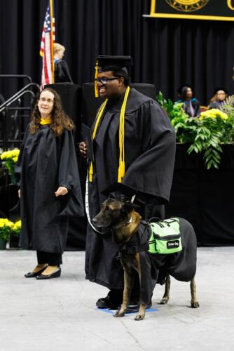 A graduate and his service dog, both dressed in caps and gowns, stand for a photo in front of the stage.