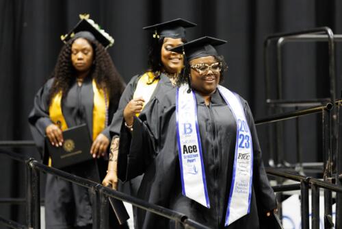 A graduate with a personalized white and blue stole walks down the ramp from the stage.
