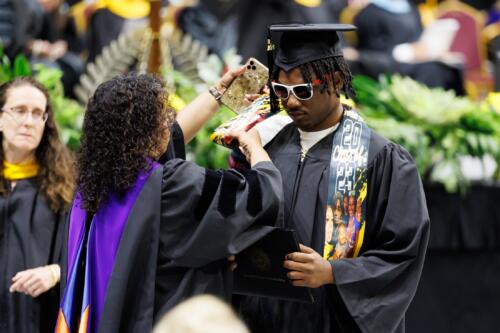 A faculty member helps a graduate wearing sunglasses straighten his personalized stole.