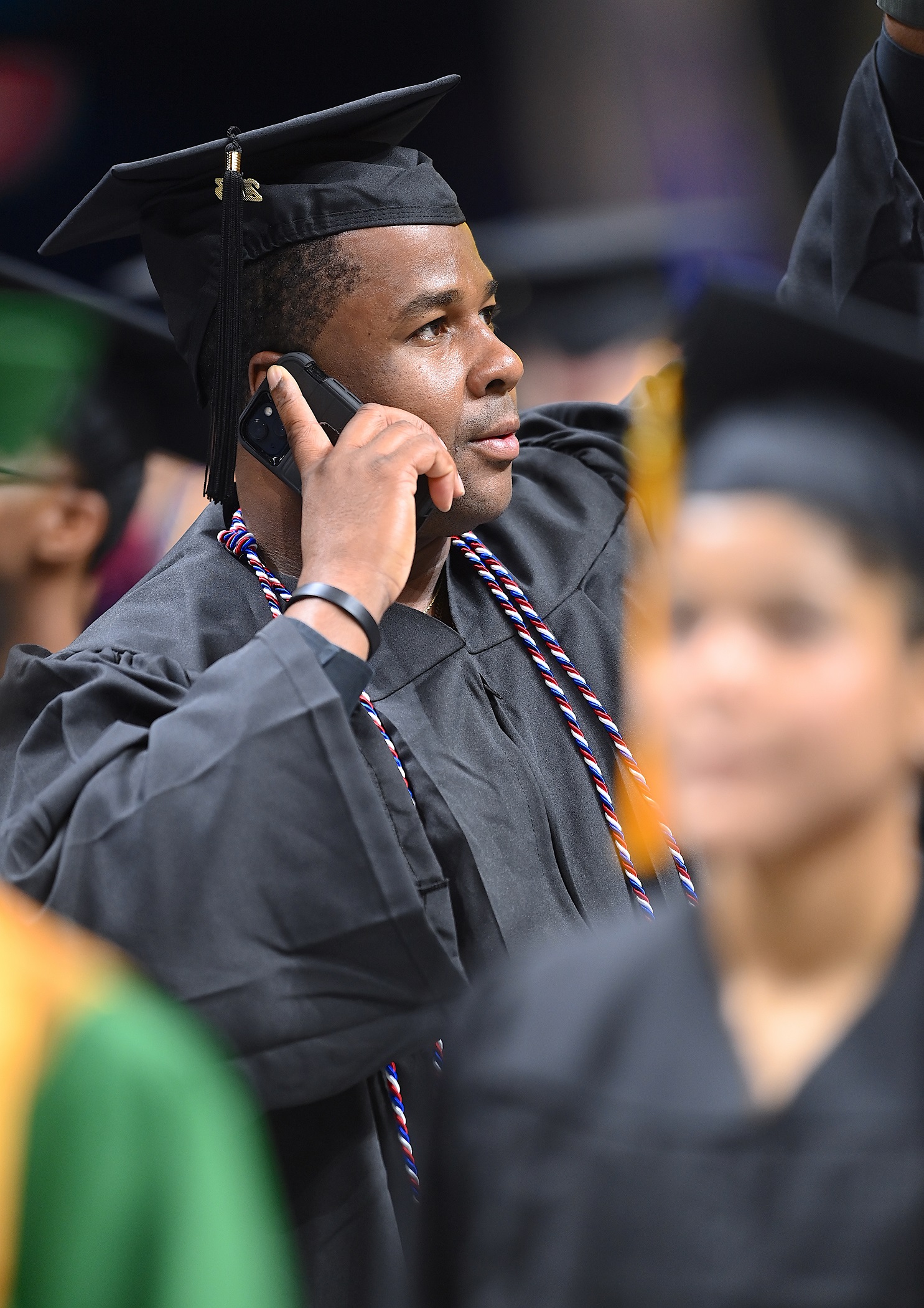A graduate wearing a red, white and blue cord, holds his phone up to his ear and raises his arm as he looks into the crowd.