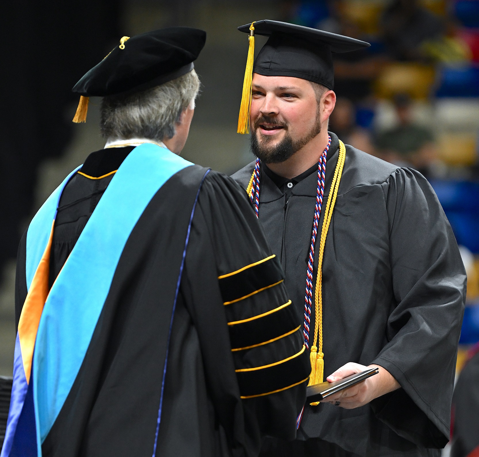 A graduate with a gold cord and a red, white and blue cord accepts his diploma folder from Dr. Mark Sorrells.