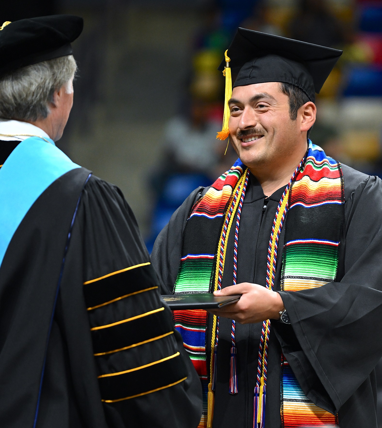 A graduate with a black moustache and a multicolored stole accepts his diploma folder from Dr. Mark Sorrells.