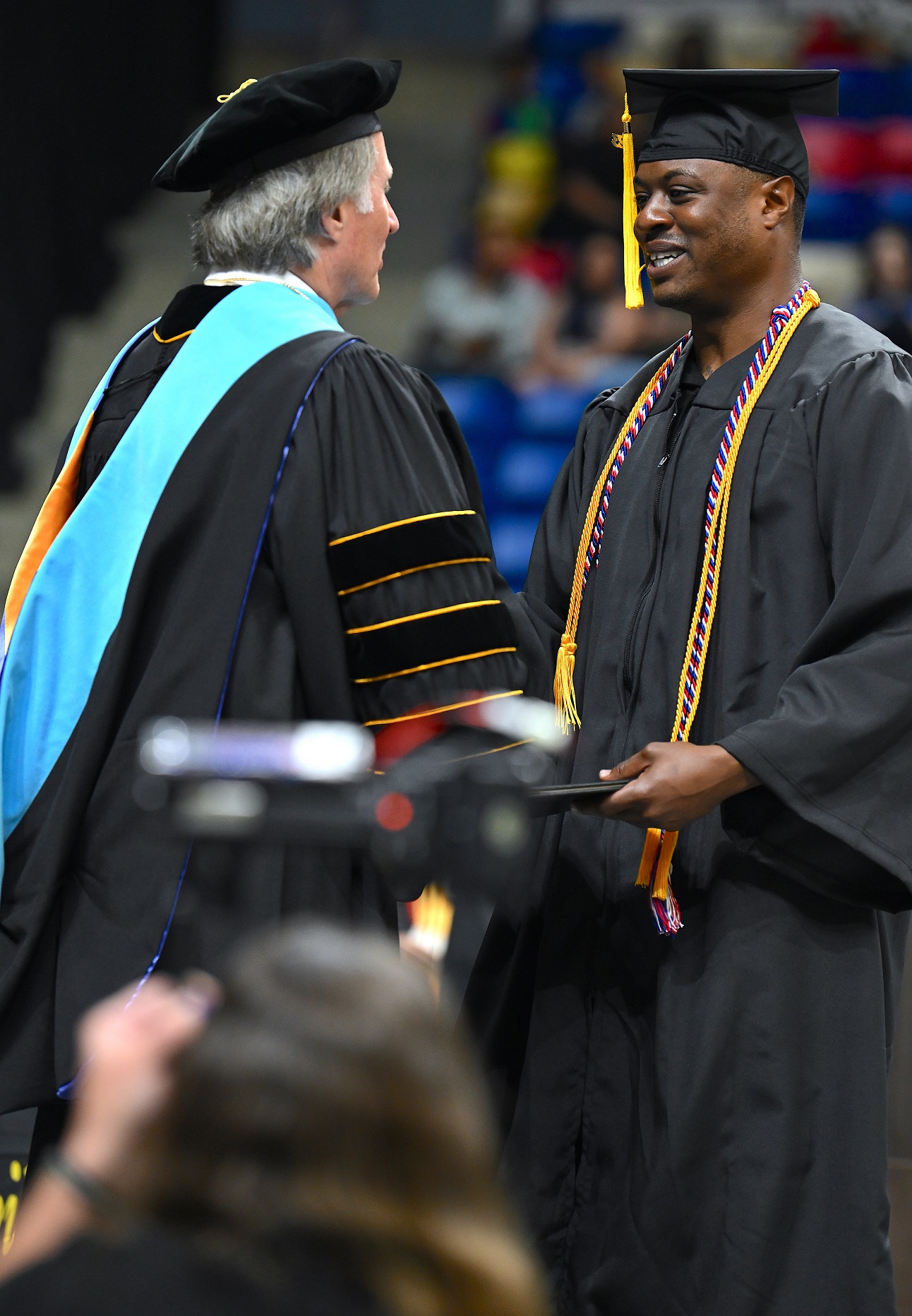 A graduate wearing a red, white and blue cord and a gold cord accepts his diploma folder from Dr. Mark Sorrells. A photographer with a large camera is visible in the foreground.