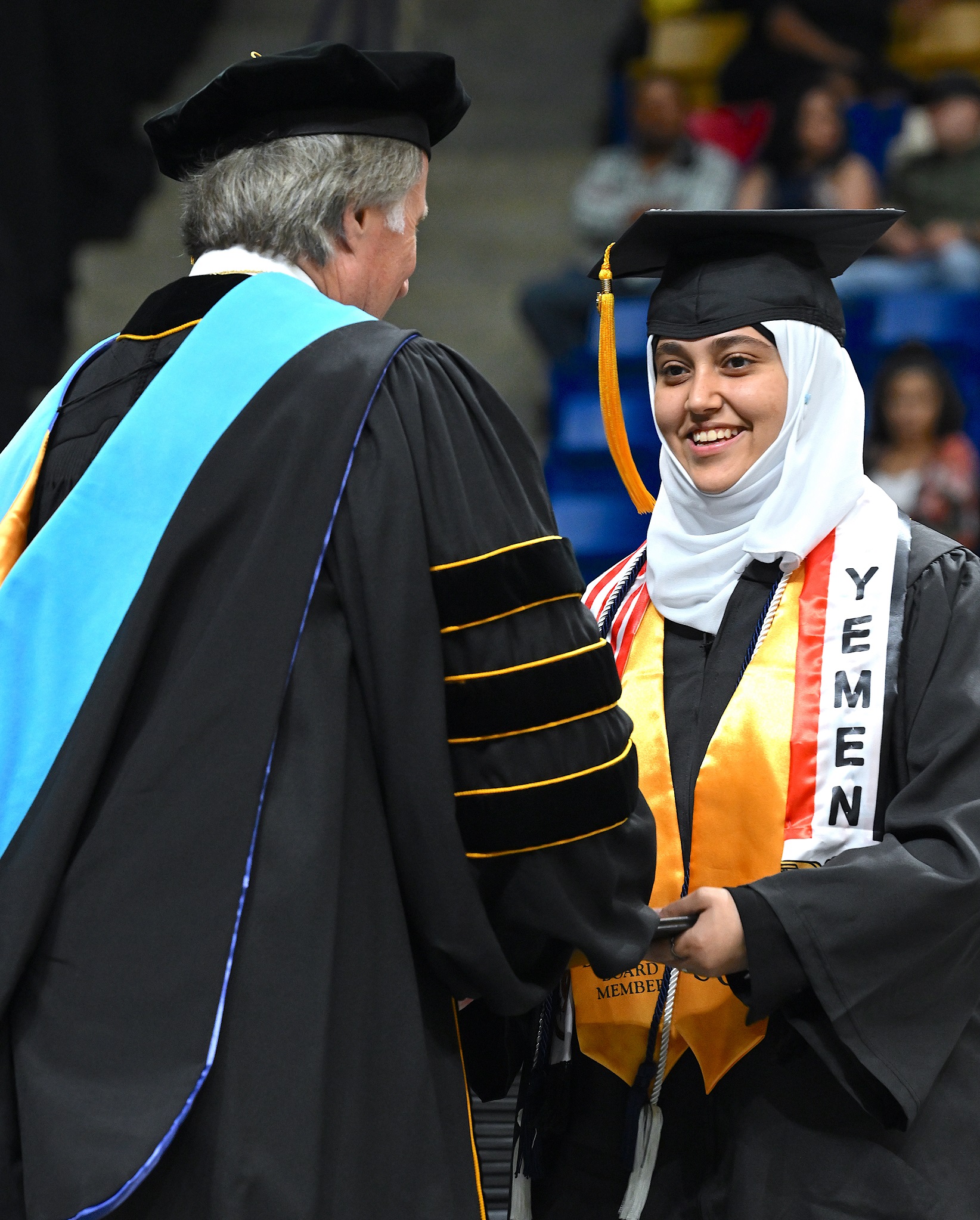 A graduate wearing a gold stole and a white stole that says "Yemen" on it smiles at Dr. Mark Sorrells. 