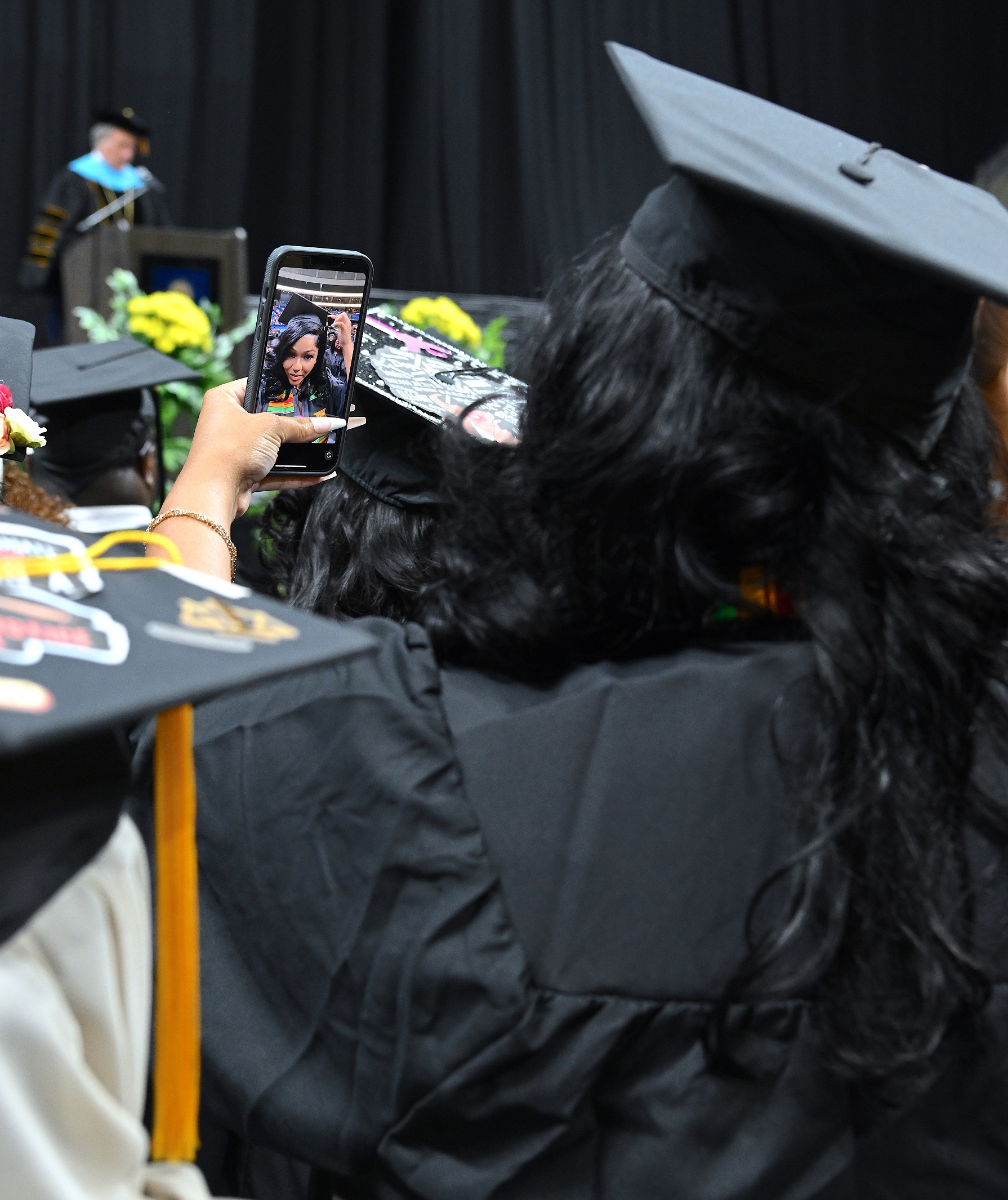A graduate, shown from behind, holds up her camera to take a selfie.