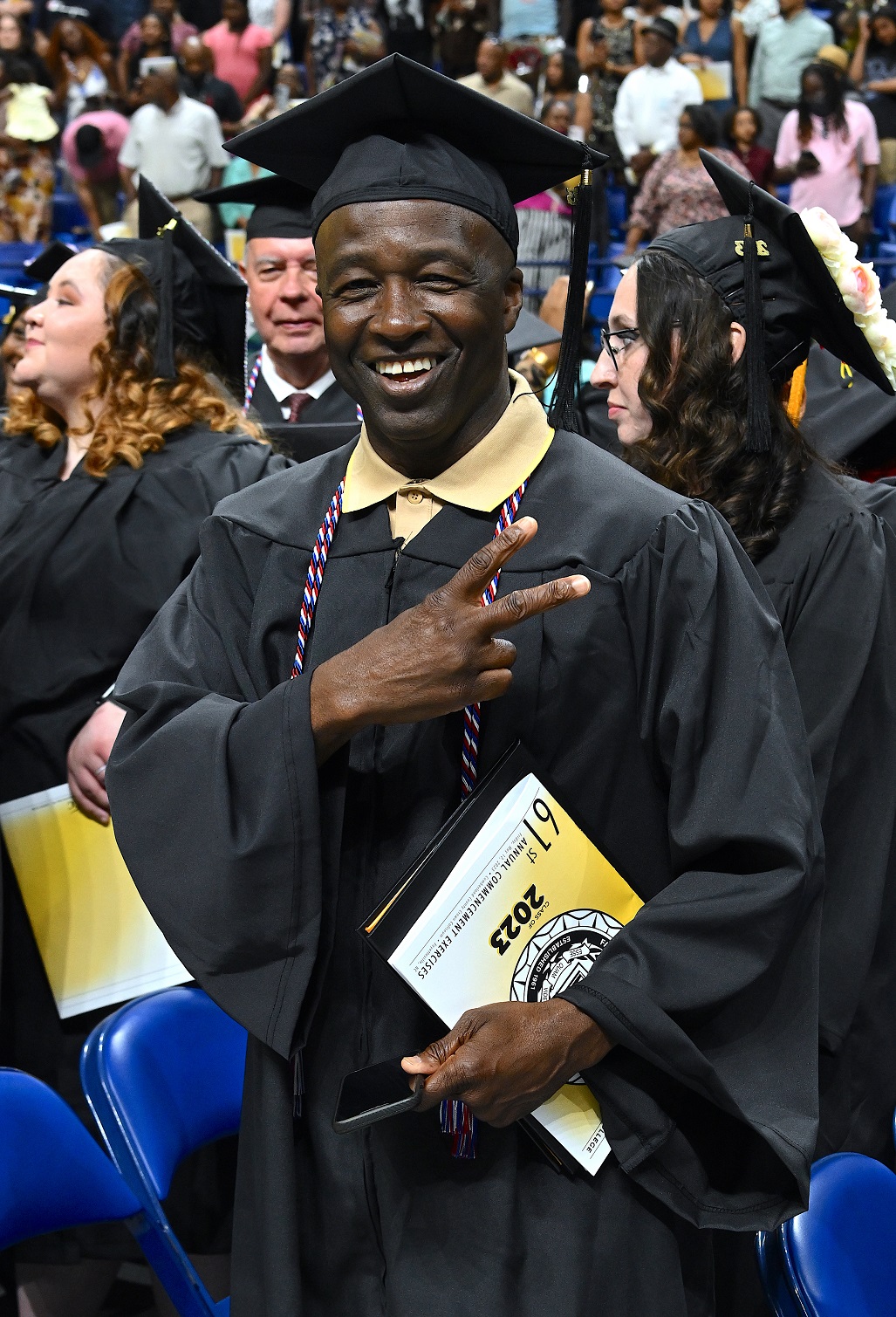 A graduate holds up two fingers over his chest while smiling at the camera.