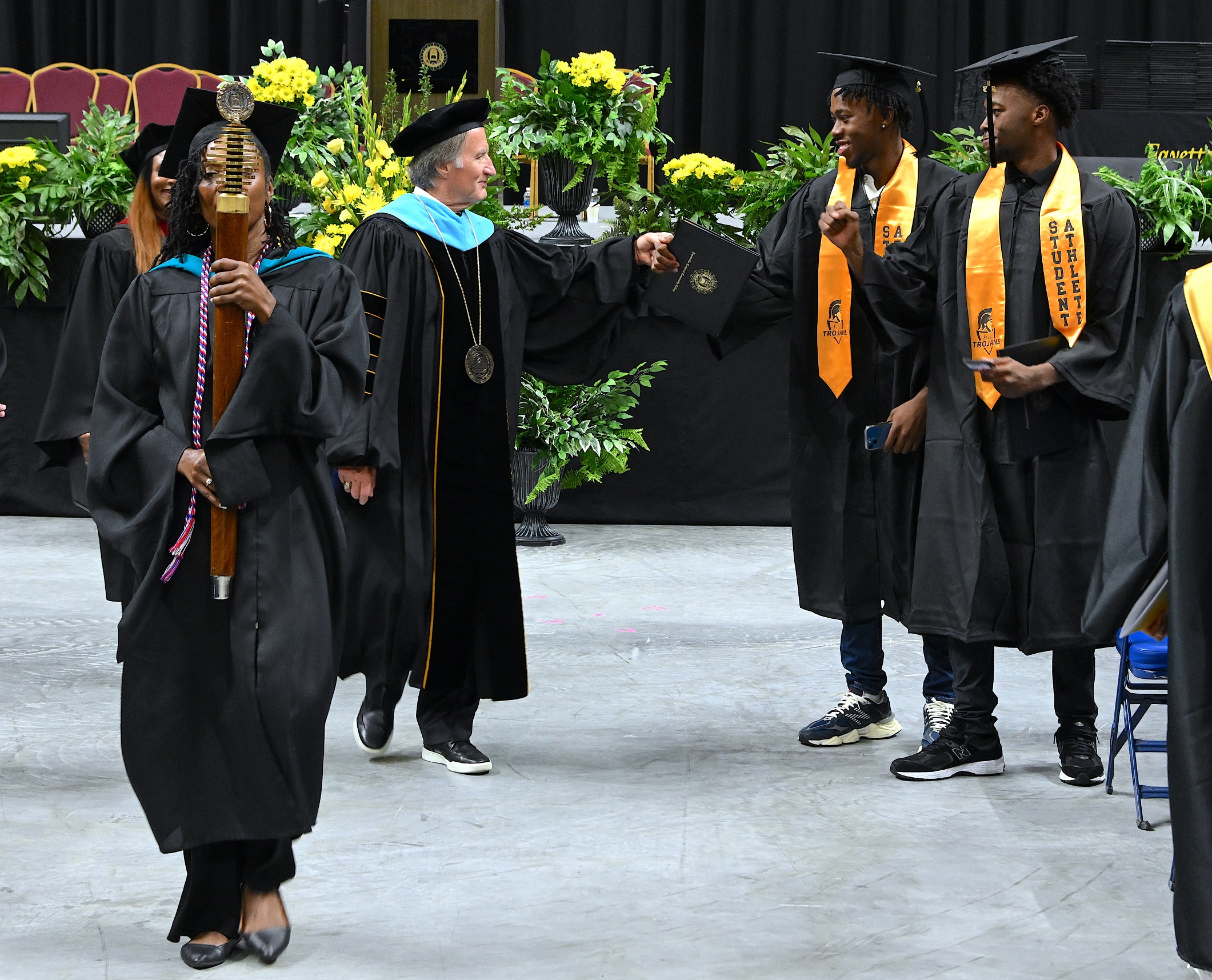 Dr. Mark Sorrells bumps fists with one of two twins who are standing at the end of a row of graduates. The FTCC Mace, being carried by Qadeerah Rasheed-Brown, is visible in the foreground.
