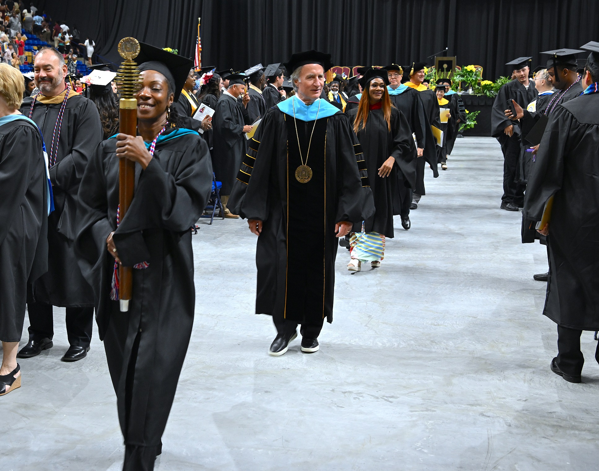 Dr. Mark Sorrells is in the center of the frame walking in the center aisle of the Crown. The FTCC Mace, being carried by Qadeerah Rasheed-Brown, is visible in the foreground.