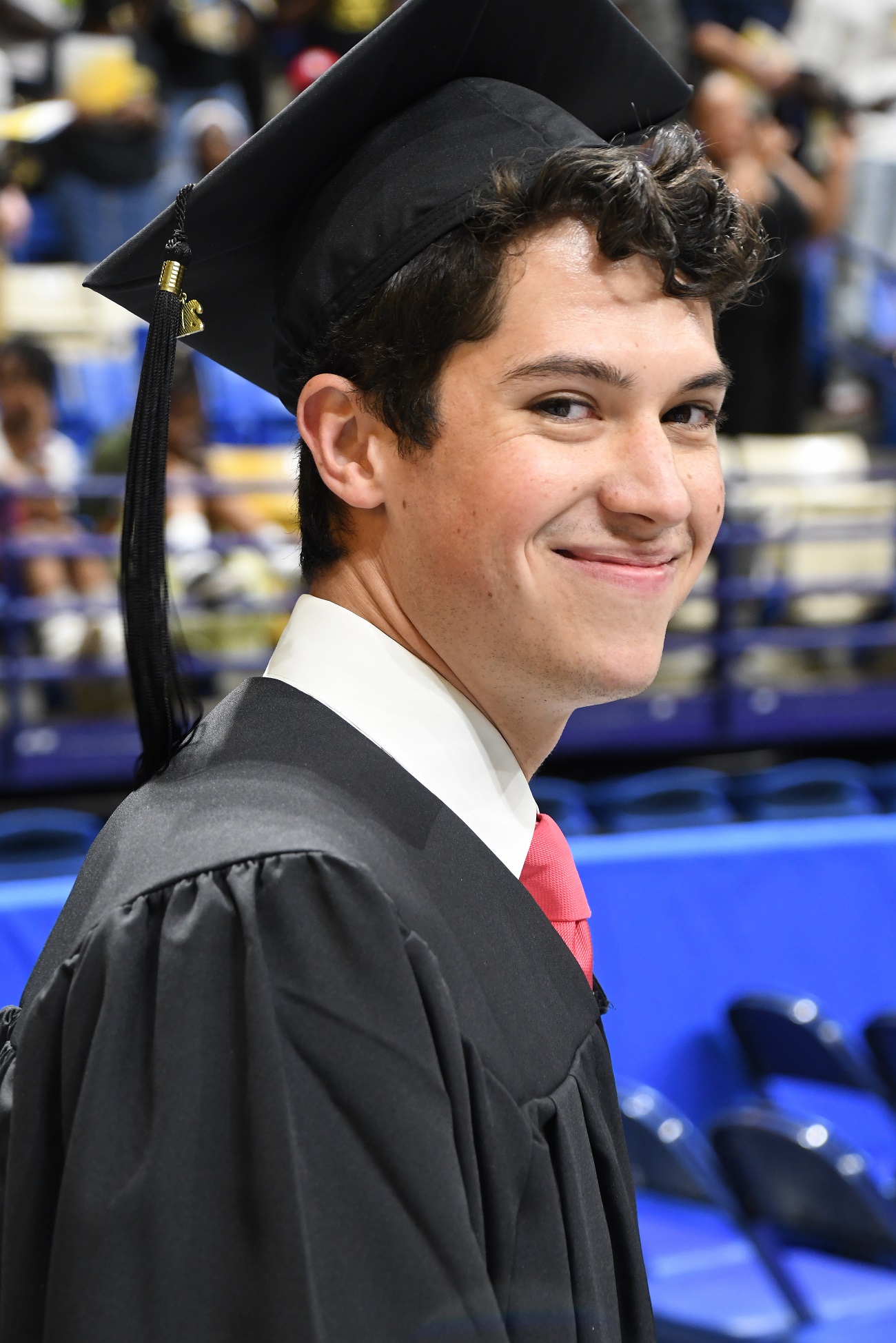 A close-up photo of a graduate smiling at the camera. He is wearing a white collar shirt and pink necktie.