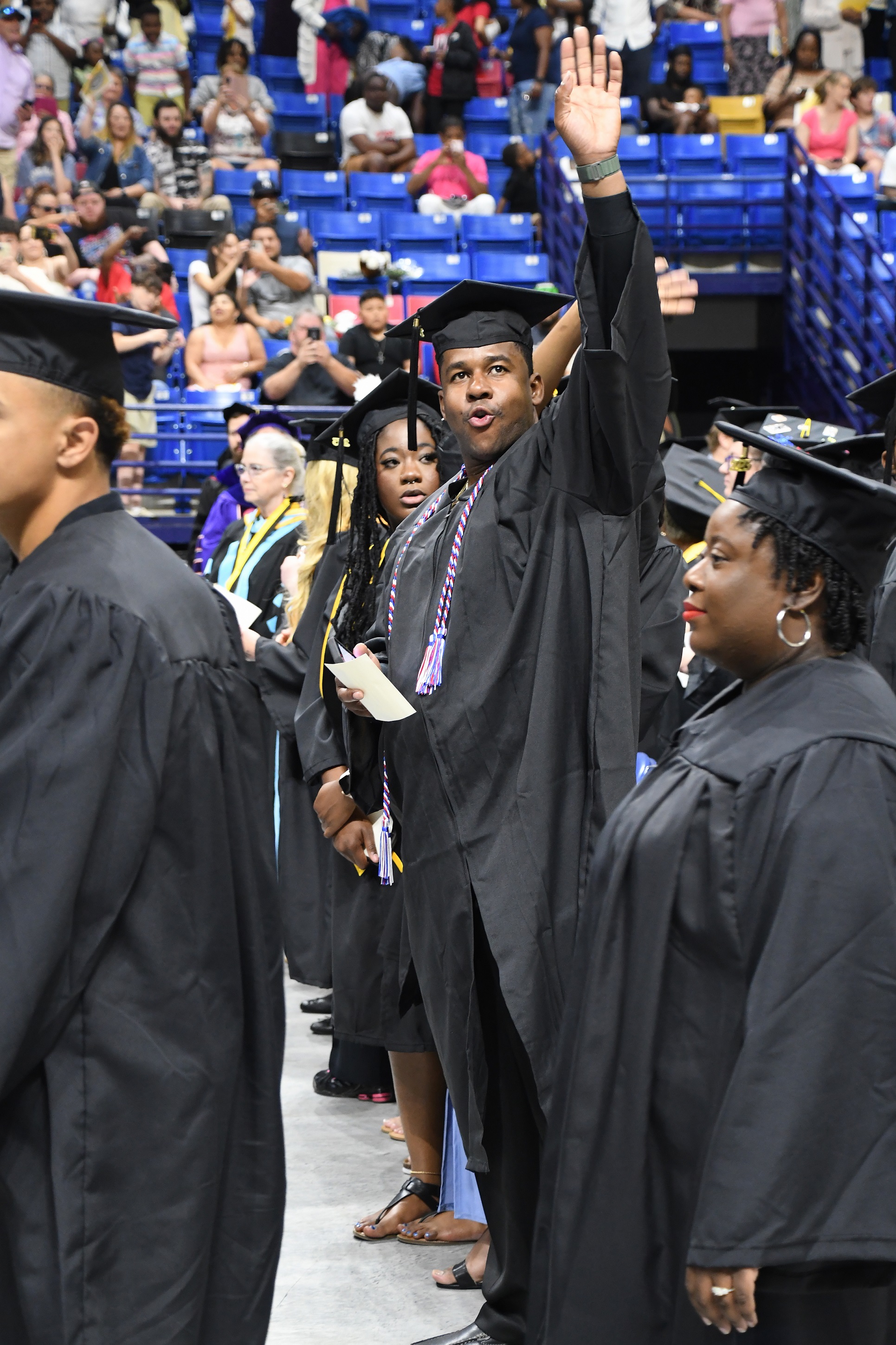A graduate standing in front of his seat in a row raises his arm to wave at the crowd.