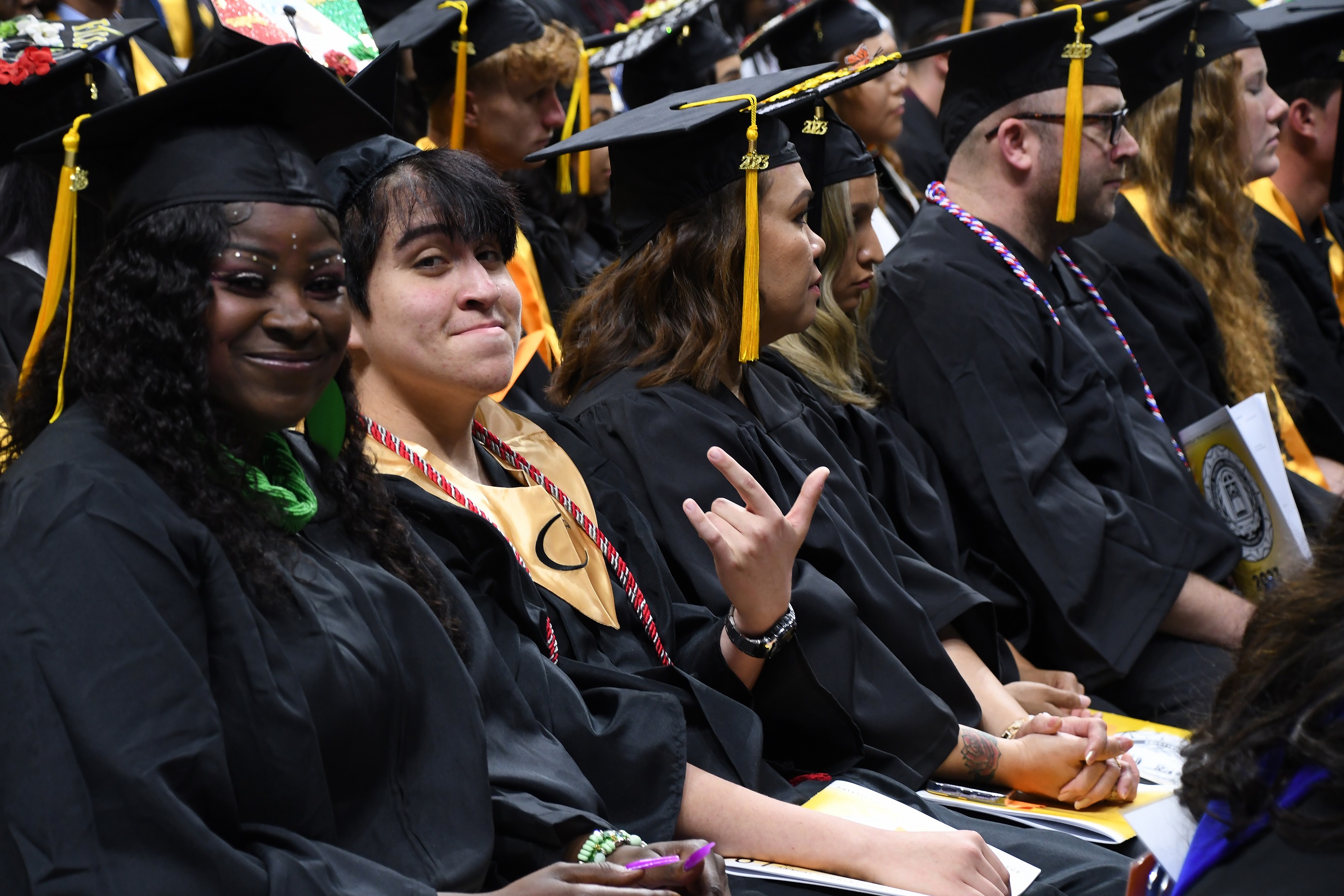 A graduate, seated in a row, holds up his index, pinky and thumb while looking at the camera.