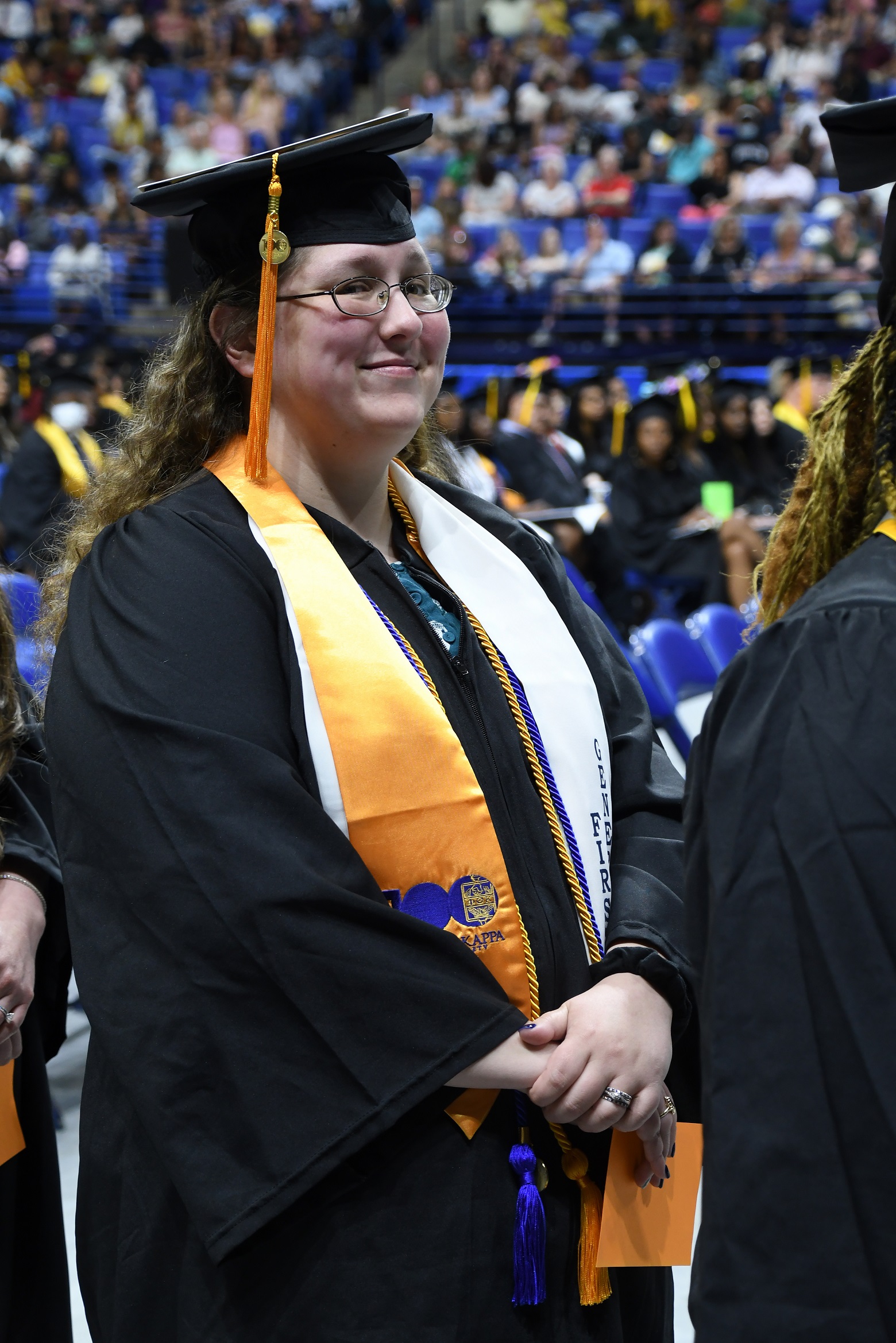 A graduate wearing a gold stole and a white stole smiles at the camera as she walks.