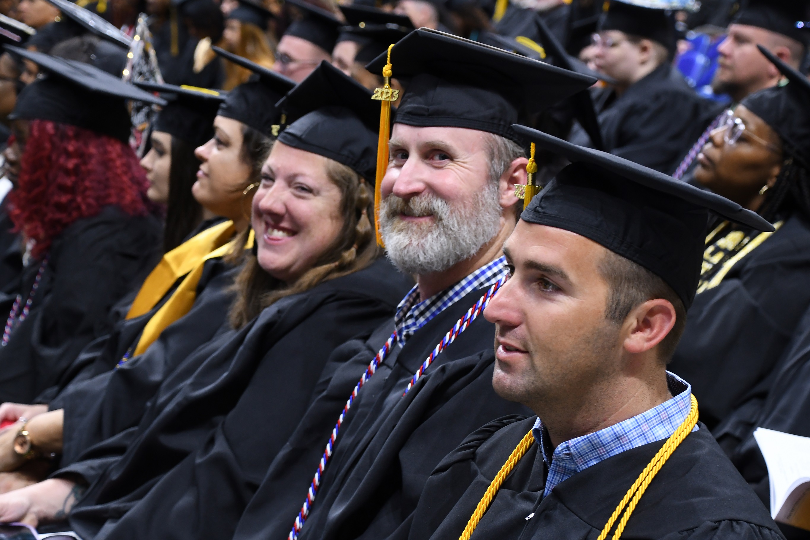 A graduate with a gray beard sits in a row and smiles at the camera.